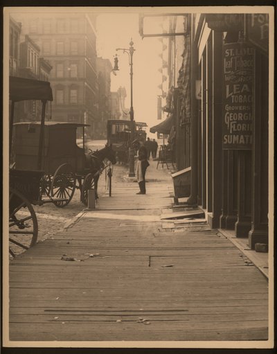 Vertical, Black and White Photograph of Horse-Drawn Vehicles Parked Against a Wood Plank Sidewalk. Storefronts Line the Sidewalk and a Man Stands on the Corner by William G. Swekosky